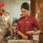 A server in a maroon uniform arranging plates, workers